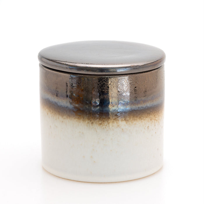 A cylindrical ceramic jar with a matching lid, featuring a glossy glaze that transitions from a dark brown and black top to a warm off-white base, set against a plain white background.
